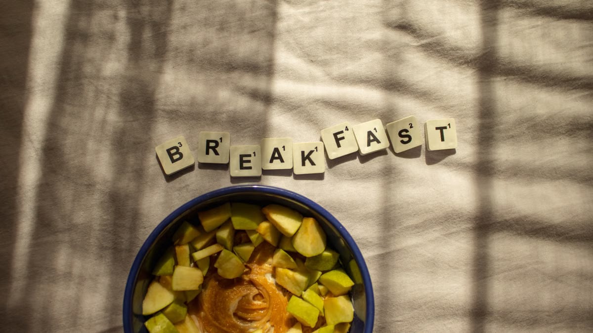Artistic breakfast scene with apple slices in a bowl and 'BREAKFAST' spelled in Scrabble tiles.